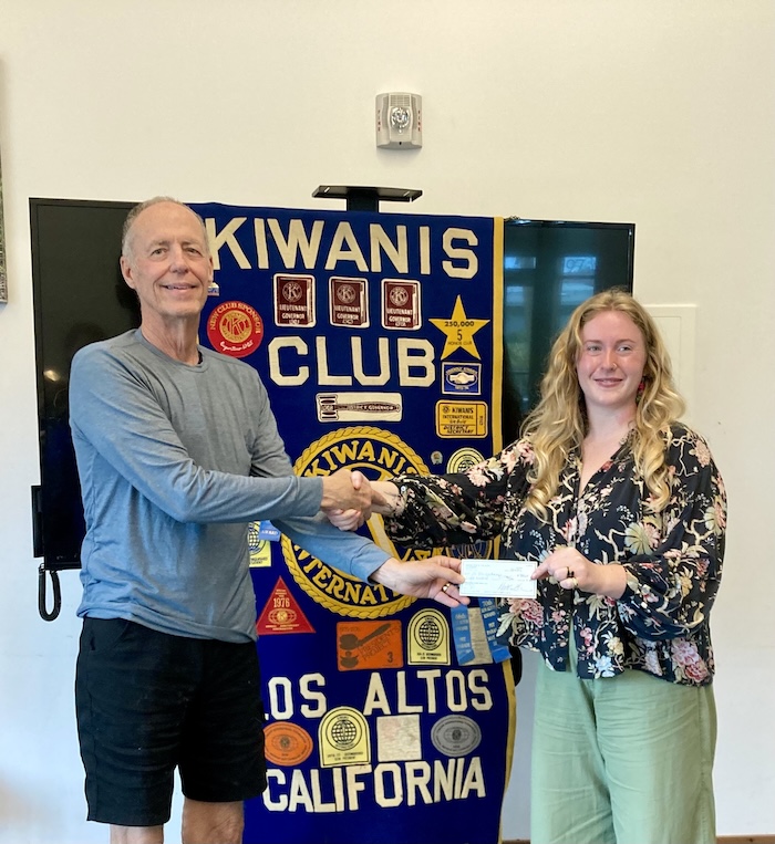Two smiling people shaking hands while one person hands a check to the other, in front of a Kiwanis Club poster